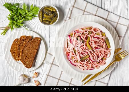 Deutscher Wurstsalat aus geschnittener lyoner-Wurst mit Gurken und Rote Zwiebel serviert auf einem weißen Teller mit goldenem Besteck Und das Roggenbrot auf dem weißen Holz Stockfoto