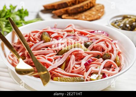 Deutscher Wurstsalat aus geschnittener lyoner-Wurst mit Gurken und Rote Zwiebel serviert auf einem weißen Teller mit goldenem Besteck Und das Roggenbrot auf dem weißen Holz Stockfoto