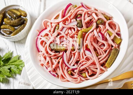 Deutscher Wurstsalat aus geschnittener lyoner-Wurst mit Gurken und Rote Zwiebel serviert auf einem weißen Teller mit goldenem Besteck Und das Roggenbrot auf dem weißen Holz Stockfoto