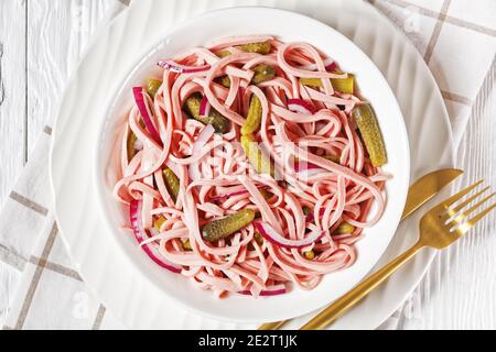 Deutscher Wurstsalat aus geschnittener lyoner-Wurst mit Gurken und Rote Zwiebel serviert auf einem weißen Teller mit goldenem Besteck Und das Roggenbrot auf dem weißen Holz Stockfoto