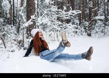 Ein glückliches Mädchen mit leuchtend roten Haaren sitzt auf dem Schnee in einem schneebedeckten Wald. Stockfoto