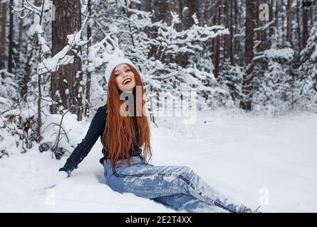 Ein glückliches Mädchen mit leuchtend roten Haaren sitzt auf dem Schnee in einem schneebedeckten Wald. Stockfoto