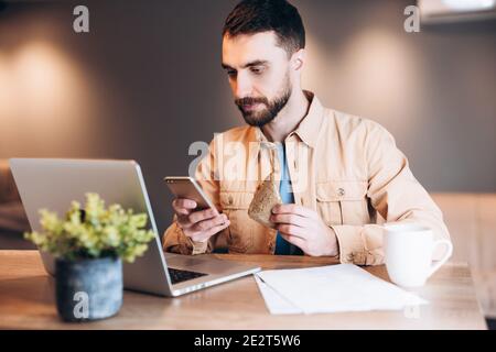 Konzentrierter Mann mit seinem Telefon und Laptop. Mann in legerer Kleidung arbeitet und isst sein Frühstück. Modernes Zimmer im Hintergrund. Telearbeiter Stockfoto
