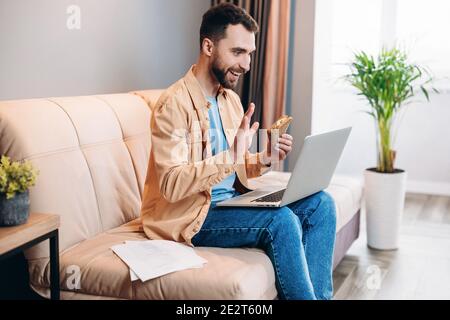 Mann spricht per Videocall, isst Sandwich und sitzt auf dem Sofa in seinem gemütlichen Wohnzimmer. Mann, der mit Freunden über seinen Laptop kommuniziert und lächelt Stockfoto