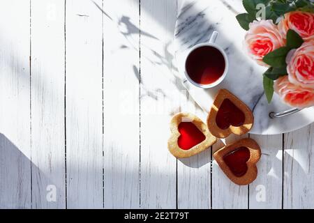 Herzförmige Kekse mit Erdbeermarmelade, Tasse Kaffee, rosa Blumen auf weißem Holztisch. Geburtstag, Valentinstag, Muttertag. Top-Ansicht. Tageslicht Stockfoto