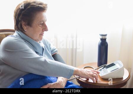 Kaukasische Frau über 70, die ihren Blutdruck zu Hause mit einem digitalen Monitor überprüft. Kurze Haare, blauer Pullover, Decke und kühles Wasser Stockfoto