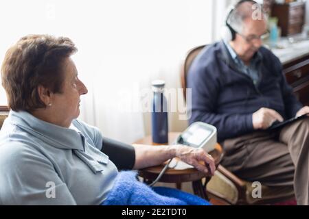 Kaukasisches Seniorenpaar zu Hause mit digitalen Geräten und Überprüfung des Blutdrucks in blauen Farben gekleidet. Stockfoto