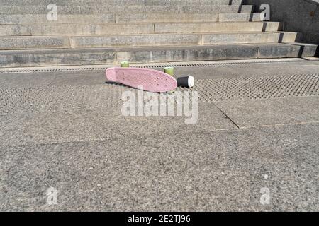 Ein pinkes Skateboard liegt an einem sonnigen Tag kopfüber unter der Treppe. Das Trauma des Straßensports und der individuellen Mobilität Stockfoto