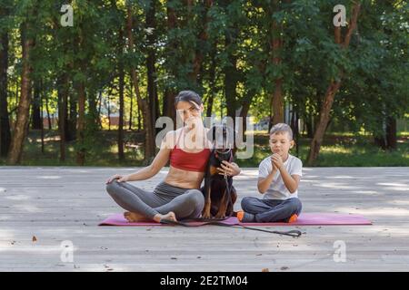 Schöne sportliche Frau umarmt niedlichen doberman Welpen bei Yoga-Klasse im Park. Kleiner Junge sitzt neben ihm und ist traurig. Das Kind ist eifersüchtig auf die mothe Stockfoto