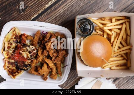 Draufsicht auf den Tisch mit Lebensmitteln in Kunststoffbehältern. Lieferung von Lebensmitteln. Hähnchensalat, Hähnchenflügel mit Sous, Burger auf pommes auf einem Holztisch. Business Lunch. Stockfoto