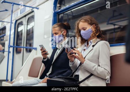 Passagiere, die Schutzmasken tragen, benutzen ihre Smartphones, während sie in einem U-Bahn-Auto sitzen. Stockfoto