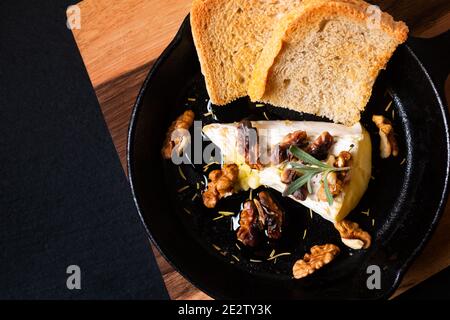 Essen Konzept Spot Fokus hausgemachte gebackenen Brie Käse mit Honig Und Walnüsse in Pfanne Eisen Pfanne auf Holzbrett mit Kopierraum aus schwarzem Schiefer Stockfoto