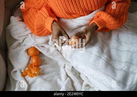 Afro Frau Hände schälen reife süße Mandarine, tragen orange Pullover, liegen im Bett unter dem Plaid Stockfoto