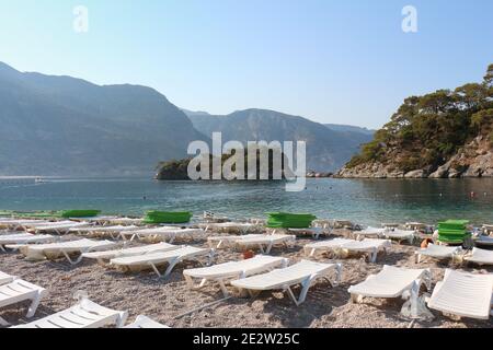 Leere Sonnenliegen und Sonnenschirme mit Blick auf türkisfarbenes Wasser und Berge Am Strand an sonnigen Tag an der blauen Lagune von Oludeniz Fethiye Türkei Stockfoto