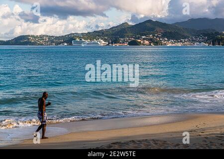 Ein Mann geht am sandigen Ufer von Grand Anse, Grenada, mit einem Getränk in der Hand entlang, während zwei Kreuzfahrtschiffe vor St. George's im Hintergrund vor Anker liegen. Stockfoto