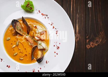 Cremige Kürbissuppe mit Meeresfrüchten, Muscheln, Garnelen, Tintenfische, auf einem weißen Teller, auf einem hölzernen Hintergrund Stockfoto