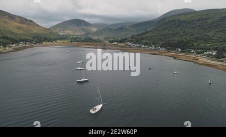 Yachten, Boote, Schiffe in Schottland Meeresbucht Antenne. Epische Berglandschaft am bewölkten Sommertag von Drohne erschossen. Filmischer Meeresurlaub an grünen Bergen an der Küste. Loch Ranza Bay, Arran Island. Stockfoto