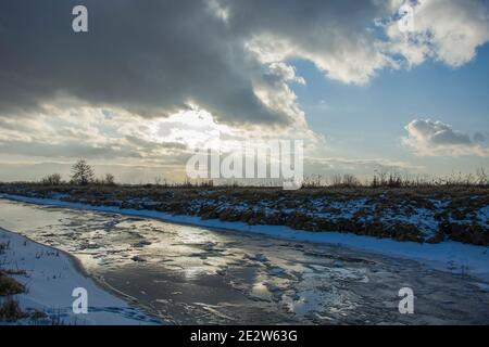 Gefrorener kleiner Fluss mit Eisscholle und Wolken mit Sonnenschein am Himmel, Winterblick Stockfoto