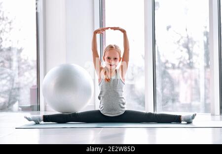 Kleine Balletttänzerin sitzt auf dem Garn mit den Händen oben im hellen Tanzsaal und macht vor dem Training Dehnen. Sportliches Kinderkonzept Stockfoto