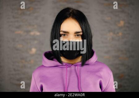 Ernsthafte junge Frau in schwarzer Schutzmaske, die Sie ansieht Über Home Wand Hintergrund Stockfoto