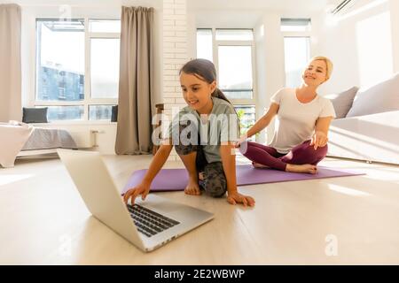 Mutter und Tochter üben online Yoga-Kurse zu Hause während der Quarantäne während der Coronavirus-Pandemie. Familie macht Sport zusammen online Stockfoto