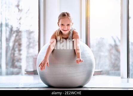 Lächelndes nettes sportliches Mädchen, das dich ansieht, während es auf dem Fitnessball im Fitnessstudio liegt. Yoga, Fitness, Pilates für Kinder Stockfoto