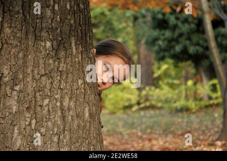 Kaukasische 8 Jahre altes Kind, Mädchen, glücklich im Park, lächelnd und versteckt sich hinter einem großen Baum Stockfoto
