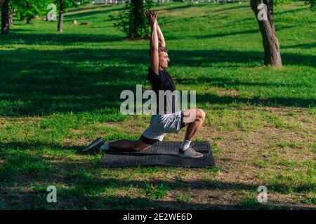 Sportlicher junger Mann, der die Beine dehnt und beim Training im Park einen Ausfallschritt nach vorne macht Stockfoto