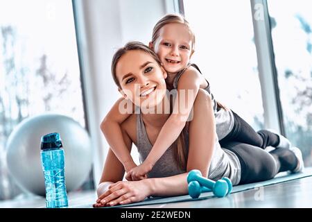 Ein kleines Mädchen liegt auf dem Rücken ihrer Mutter und ruht sich nach dem Training in Fitnessatmosphäre mit Hanteln, Matte, Fitnessball, Flasche Wasser auf dem Boden aus. Stockfoto