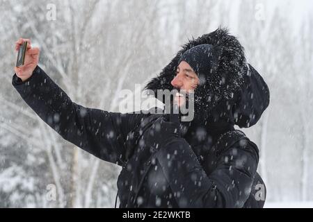 Hipster Mann mit einem Bart nimmt ein Selfie mit seinem Mobile mit einem Hut und Handschuhe, um sich vor zu schützen Die Kälte, während ein schwerer Schnee fällt in einem Par Stockfoto