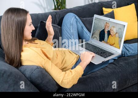 Porträt einer jungen Frau mit Laptop, spricht sie online mit Großeltern, während sie zu Hause auf dem Sofa liegt, Senioren auf dem Bildschirm. Konzept von Stockfoto