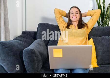 Freiberuflerin Erholung nach harten Arbeitstag. Eine Frau in Freizeitkleidung mit Laptop auf der Couch lehnte sich zurück wirft die Hände hinter den Kopf, macht eine Pause Stockfoto
