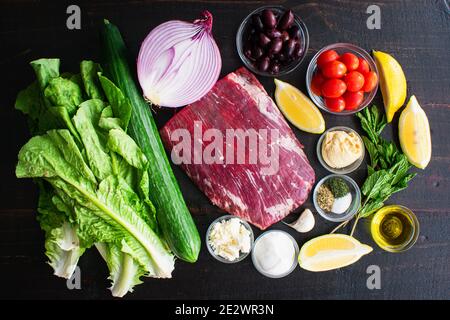 Mediterrane Steakschüssel Zutaten: Rohes Flankensteak, Obst, Gemüse und Kräuter, die für die Salatzumischung auf dunklem Holz verwendet werden Stockfoto
