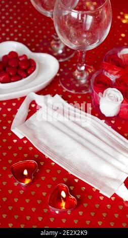 Valentinstag Konzepttisch. Romantisches Abendessen mit medizinischer Maske. Coronavirus 2021. Gesichtsschutzmaske und rote Tischeinstellung. Vertikales Foto Stockfoto