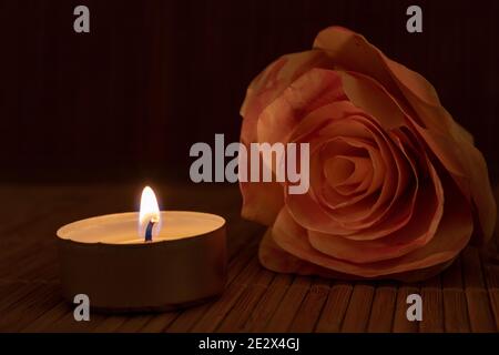 Rose und brennende Kerze in der Dunkelheit mit Kopierraum. Nahaufnahme von brennendem Teelicht und Rose auf Holztisch vor dunklem Hintergrund. Stockfoto