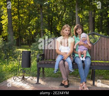 Zwei Freunde mit Kindern ruhen im Sommer im Park. Sitzen auf einer Bank. Stockfoto