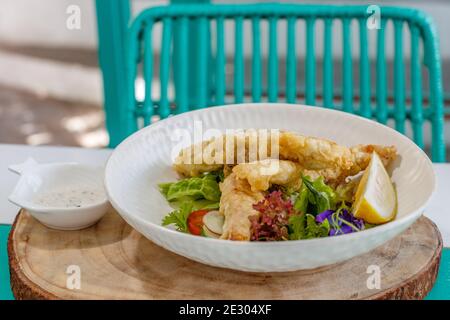 Gebutterter Fisch und grüner Salat mit Sauce und Zitrone, verziert mit Schmetterlingsblüten. Serviert auf einer weißen Keramikplatte. Stockfoto