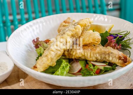 Gebutterter Fisch und grüner Salat mit Sauce und Zitrone, verziert mit Schmetterlingsblüten. Serviert auf einer weißen Keramikplatte. Stockfoto