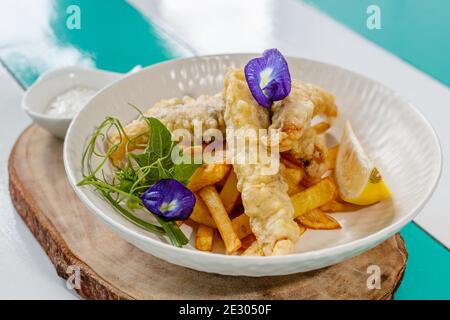 Fisch und Chips mit Sauce und Zitrone, verziert mit Schmetterlingsblüten. Serviert auf einer weißen Keramikplatte. Stockfoto
