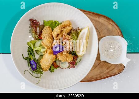 Gebutterter Fisch und grüner Salat mit Sauce und Zitrone, verziert mit Schmetterlingsblüten. Serviert auf einer weißen Keramikplatte. Stockfoto