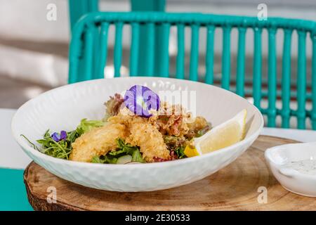 Gebutterter Fisch und grüner Salat mit Sauce und Zitrone, verziert mit Schmetterlingsblüten. Serviert auf einer weißen Keramikplatte. Stockfoto