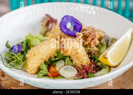 Gebutterter Fisch und grüner Salat mit Sauce und Zitrone, verziert mit Schmetterlingsblüten. Serviert auf einer weißen Keramikplatte. Stockfoto