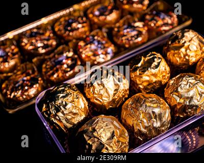 Eine Schachtel mit runden Bonbons in goldener Folie auf schwarzem Hintergrund. Verpackte Pralinen. Runder Kuchen. Süßes Essen. Süßes Geschenk. Goldene Farbe. Food-Foto. Produkt Stockfoto