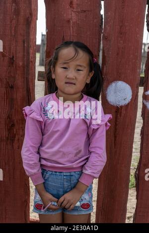 Olgii, Mongolei - 7. August 2019: Mongolisches junges Mädchen mit violettem Pullover vor einem roten Zaun. Stockfoto