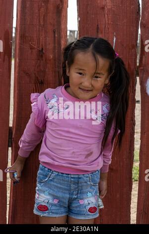 Olgii, Mongolei - 7. August 2019: Mongolisches Mädchen lächelnd und mit violettem Pullover vor einem roten Zaun. Stockfoto