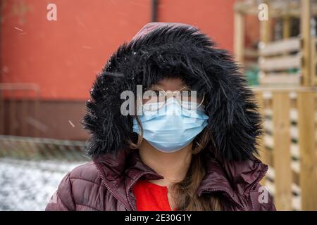 Ein Teenager-Mädchen mit in einer Winterjacke mit einer Kapuze trägt eine schützende Gesichtsmaske. Bilder bei kalten Temperaturen und Schneebedingungen Stockfoto