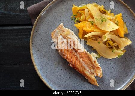 Kabeljaufisch mit Fenchelgemüse und orangefarbenen Scheiben auf einem blauen Teller und einem dunklen Tisch, hohe Ansicht von oben, ausgewählter Fokus Stockfoto