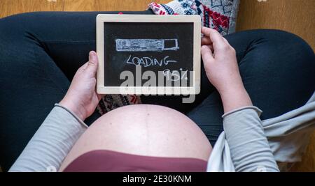 Draufsicht auf eine schwangere Frau, die mit gekreuzten Beinen auf dem Boden sitzt und eine kleine Tafel hält Stockfoto