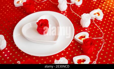 Ort Einstellung in rot und weiß - für Valentinstag oder andere Veranstaltung. Weißer Teller in Herzform mit einem Dekor aus Rosen auf rotem Hintergrund Stockfoto