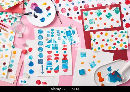 Draufsicht auf Platten mit Flecken von bunten Farbe platziert Auf rosafarbenem Hintergrund inmitten von Papierblättern mit abstrakter Geometrie Muster Stockfoto